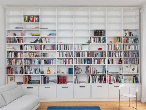 Maßgefertigtes weißes Regal, das eine komplette Wand eines Wohnzimmers einnimmt. Offene Fächer, Schubladen und dezente Beleuchtung bieten viel Stauraum für Bücher und Dekoration.