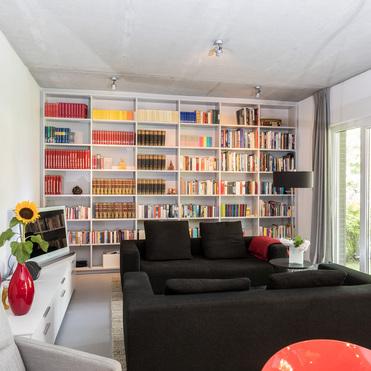 Großes weißes Bücherregal von ASADA – Stilvolle Wohnraumgestaltung Maßgefertigtes weißes Regal im Wohnzimmer, das eine komplette Wand einnimmt. Die Kombination aus farblich sortierten Büchern und eleganter Einrichtung schafft eine gemütliche Atmosphäre.