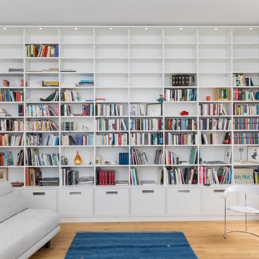 Großes weißes Bücherregal mit Beleuchtung von ASADA – Stilvolle Aufbewahrungslösung Maßgefertigtes weißes Regal, das eine komplette Wand eines Wohnzimmers einnimmt. Offene Fächer, Schubladen und dezente Beleuchtung bieten viel Stauraum für Bücher und Dekoration.