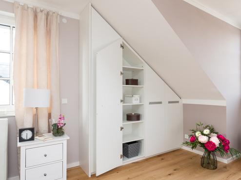 Ma&szlig;gefertigter Einbauschrank unter einer Dachschr&auml;ge im Schlafzimmer. Die wei&szlig;en T&uuml;ren mit integrierten Griffen verbergen Regalf&auml;cher, die Platz f&uuml;r Boxen und Aufbewahrungsbeh&auml;lter bieten.