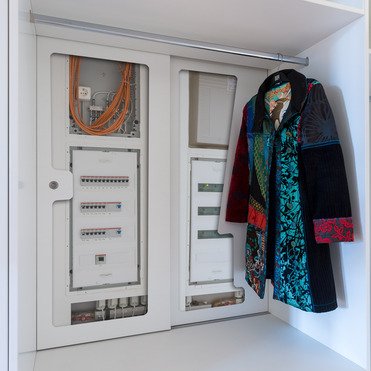 Ein maßgeschneiderter Schrank, der Technikkomponenten wie Sicherungskästen und Kabel sauber integriert, während daneben eine bunte Jacke hängt. Der Schrank bietet praktischen Stauraum.