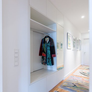 Ein heller Flur mit einem maßgefertigten Einbauschrank, der eine bunte Jacke präsentiert. Der Flur ist mit Kunstwerken an der Wand und farbenfrohen Teppichen dekoriert.