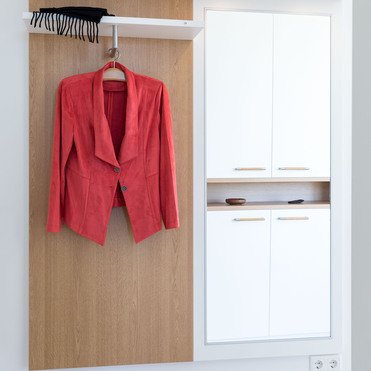 Eine maßgefertigte Garderobe aus Holz und weißem Material mit beleuchtetem Ablagefach, an der eine rote Jacke hängt. Ein Regal bietet zusätzlichen Stauraum für kleine Gegenstände.