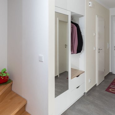 Ein moderner Eingangsbereich mit einer maßgefertigten Garderobe, die einen Spiegel, eine Sitzbank mit Stauraum und Kleiderhaken enthält. Im Hintergrund ist eine dekorierte Eingangstür zu sehen.