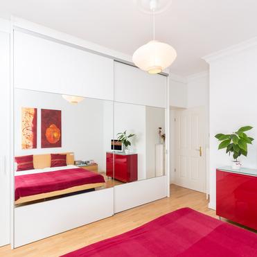 Helles Schlafzimmer mit glänzendem Kleiderschrank nach Maß, der als Spiegel fungiert, roten Akzenten und minimalistischer Einrichtung.