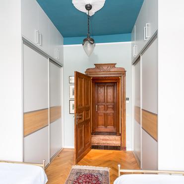 Zwei weiße, maßgefertigte Kleiderschränke flankieren eine Holztür, über einem persischen Teppich, unter einer blauen Decke.