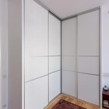 Eckiger Kleiderschrank nach Maß mit glatten weißen Türen auf Parkettboden neben einem orientalischen Teppich.