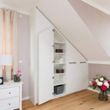 Ein weißer, unter einer Dachschräge maßgefertigter Kleiderschrank, in einem Raum mit Holzboden und frischen Blumen.
