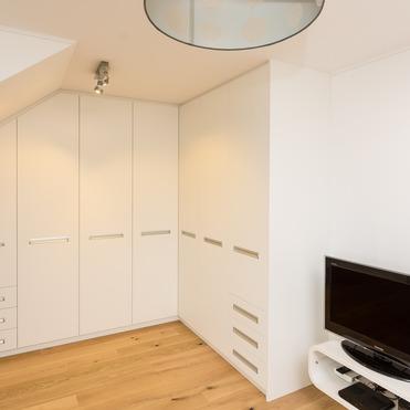 Ein helles Zimmer mit Holzboden und einem großen, weißen Kleiderschrank nach Maß, eingepasst unter eine Dachschräge.