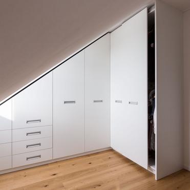 Ein moderner, weißer Kleiderschrank nach Maß, eingepasst unter eine Dachschräge, mit Kombination aus Schubladen und Schiebetüren, auf einem hellen Holzboden.