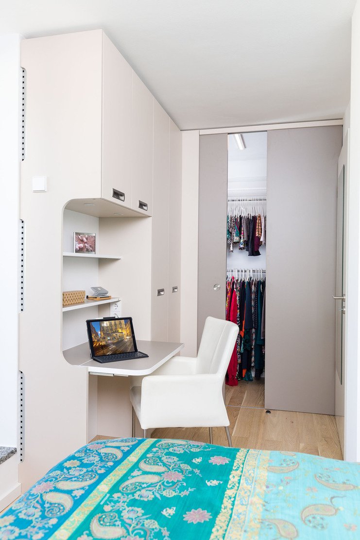 Kompakter Arbeitsbereich mit integriertem Kleiderschrank Ein moderner Arbeitsbereich in einer Ecke mit einem gebogenen weißen Schreibtisch, einem Laptop darauf und einem weißen Stuhl. Im Hintergrund ist der Eingang zu einem integrierten Kleiderschrank sichtbar.