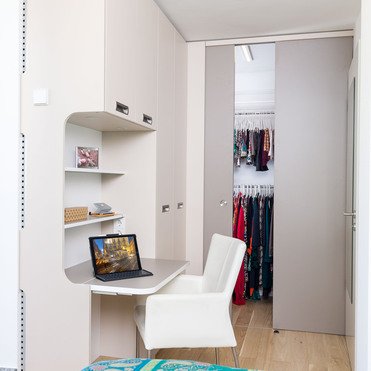 Ein moderner Arbeitsbereich in einer Ecke mit einem gebogenen weißen Schreibtisch, einem Laptop darauf und einem weißen Stuhl. Im Hintergrund ist der Eingang zu einem integrierten Kleiderschrank sichtbar.