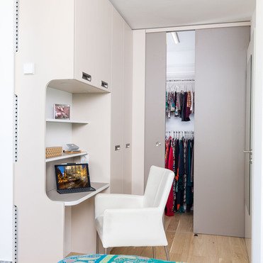 Ein maßgeschneiderter Arbeitsbereich mit einem runden Schreibtisch und einem weißen Stuhl, links davon eine offene Tür zum Kleiderschrank, gefüllt mit farbenfroher Kleidung.
