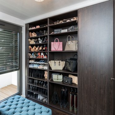 Ein luxuriöser Ankleideraum mit einem großen, dunkelholzfarbenen Kleiderschrank nach Maß, der Schuhe und Taschen ordentlich präsentiert. Im Vordergrund eine große, blaue Samtbank.