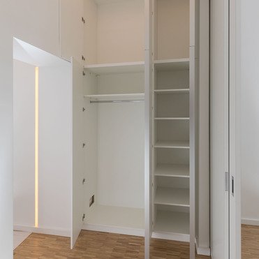 Ein leerer, modern gestalteter begehbarer Kleiderschrank in Weiß mit maßgeschneiderten Regalen und Kleiderstangen, beleuchtet von einer schmalen, vertikalen Lichtleiste.
