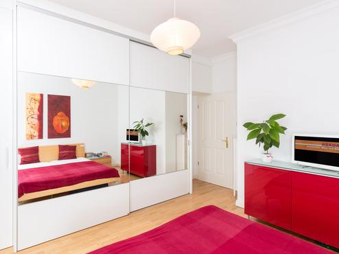 Helles Schlafzimmer mit glänzendem Kleiderschrank nach Maß, der als Spiegel fungiert, roten Akzenten und minimalistischer Einrichtung.
