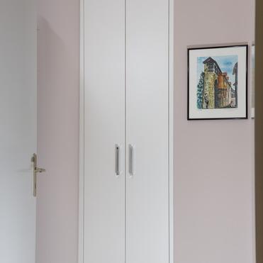 Ein schlichter, weißer Nischenmöbel nach Maß, eingepasst in eine kleine Raumecke, neben einem Bild an der Wand und einer Tür mit Glasfenster.