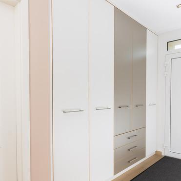 Ein großer weißer Einbauschrank mit schlichten Griffen und dezenter kupferfarbener Akzentlinie, integriert in eine helle Flurumgebung neben einer Tür mit Spiegel.