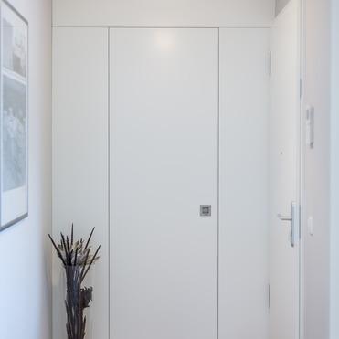 Ein schlicht gestalteter Flur mit einem nahtlos integrierten, weißen Schrank, neben einer modernen Tür und einem dekorativen Ast in einer Glasvase.