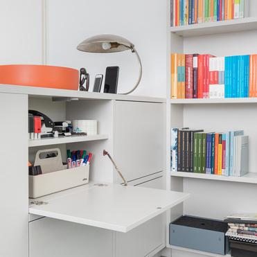 Ein weißes Sideboard mit offener Klappe zeigt Büroartikel wie Stifte und Heftklammern. Daneben steht ein Bücherregal mit farbenfrohen Büchern. Eine Tischlampe und Deko-Objekte sind ebenfalls sichtbar.