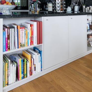 Ein weißes Sideboard in einer Küche, gefüllt mit Kochbüchern und dekorativen Elementen. Auf der schwarzen Arbeitsplatte stehen eine Obstschale, Gewürze und Küchenutensilien. Holzboden im Vordergrund.