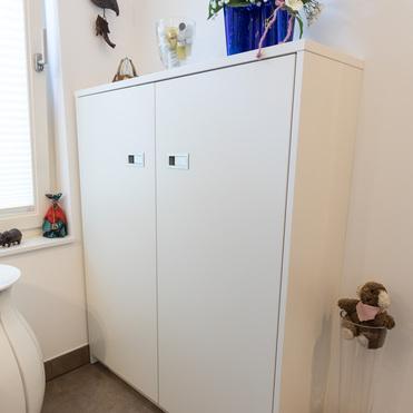 Ein weißes Sideboard mit zwei Türen, darauf eine Vase mit lila Blumen. An der Wand hängen dekorative Vögel. Neben dem Sideboard stehen kleine Dekofiguren und ein Stofftier in einem Glasgefäß.