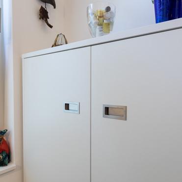 Nahaufnahme eines weißen Sideboards mit eingelassenen Griffen. Auf der Oberfläche stehen dekorative Objekte, darunter eine Glasvase mit kleinen Flaschen. Links ist ein Fenster mit einer Figur auf der Fensterbank zu sehen.