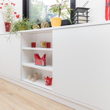 Modernes, weißes Sideboard mit offenem Regal und dekorativen Elementen wie Pflanzen und Kerzen, unter einem Fenster, das natürliches Licht in den Raum bringt.