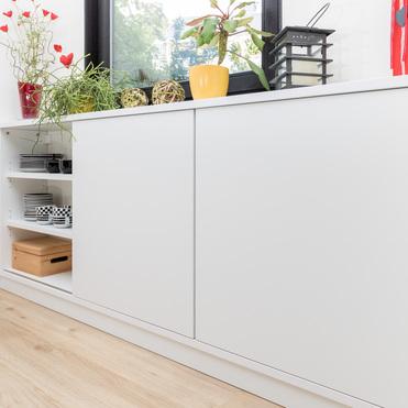 Modernes, weißes Sideboard mit offenem Regal und dekorativen Elementen wie Pflanzen und Kerzen, unter einem Fenster, das natürliches Licht in den Raum bringt.