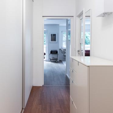 Ein schlichter, moderner Flur mit Holzfußboden und weißen Wänden. Rechts steht ein weißes Sideboard, links ist eine Schiebetür. Im Hintergrund ist ein helles Wohnzimmer zu sehen.