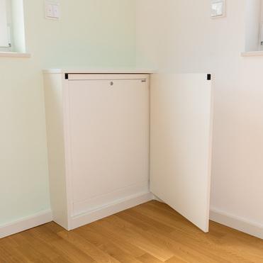 Ein weißes Sideboard mit einer offenen Tür steht in einer Ecke eines Raumes mit hellem Holzfußboden. Das Sideboard befindet sich zwischen zwei Fenstern, die etwas von der Wand zu sehen sind.