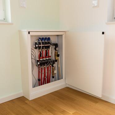 Ein weißes Sideboard in einer Ecke eines Raumes mit offenem Fach, das Heizungsrohre, Ventile und Messgeräte zeigt. Das Sideboard steht zwischen zwei Fenstern, mit hellem Holzfußboden darunter.