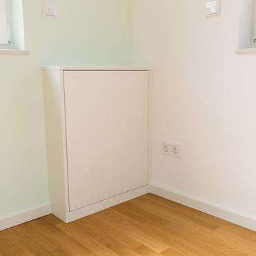 Ein weißes, geschlossenes Sideboard steht in einer Ecke eines Raumes mit hellem Holzfußboden. Das Sideboard befindet sich zwischen zwei Fenstern, und eine Steckdose ist an der Wand daneben sichtbar.