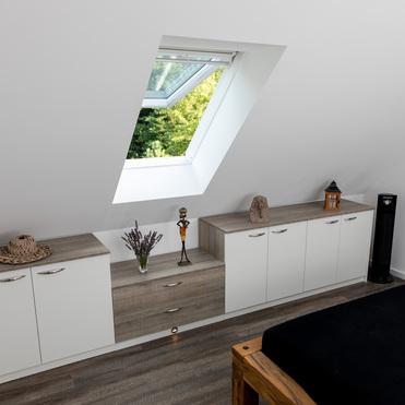 Ein Schlafzimmer mit schräger Decke und Dachfenster. Unter dem Fenster stehen mehrere Sideboards in Holzoptik mit Dekorationen, darunter Figuren und eine Laterne. Ein Bett ist teilweise sichtbar.