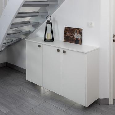 Ein weißes Sideboard steht unter einer Treppe mit grauen Stufen. Auf dem Sideboard befinden sich eine Laterne und ein Bild mit Schokoladenmotiv. Der Boden ist mit grauen Fliesen ausgelegt.