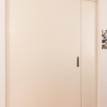 Eine geschlossene, cremefarbene Zimmertür mit einem schlichten Griff in einem Raum mit hellem Holzfußboden. Rechts an der Wand sind mehrere Fotos angebracht.