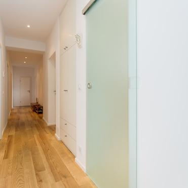 Ein heller Flur mit Holzfußboden und einer matten Glasschiebetür. An der linken Seite befinden sich Wandschränke, und der Flur führt zu einer weißen Zimmertür im Hintergrund. Die Wände sind weiß gestrichen.