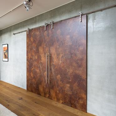 Zwei große, rostfarbene Schiebetüren aus Metall mit langen Griffen in einem modernen Raum mit Betonwänden und Holzboden. Die Türen laufen an einer Schiene entlang.