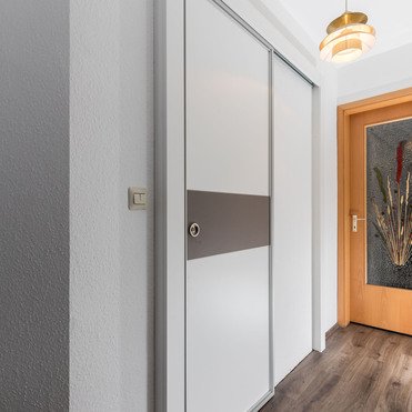 Eine moderne Schiebetüranlage aus Glas, teilweise mattiert, in einem Flur mit grauem Holzfußboden. Die Schiebetür ist abschließbar und trennt den Flur vom Badezimmer. Im Hintergrund ist eine Holztür mit Glas- und Blumenmuster zu