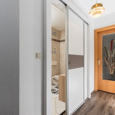 Eine geöffnete Schiebetüranlage aus Glas, teilweise mattiert, in einem Flur mit grauem Holzfußboden. Durch die geöffnete Tür ist ein Badezimmer mit beigen Fliesen und einer Toilette zu sehen. Im Hintergrund ist eine Holztür